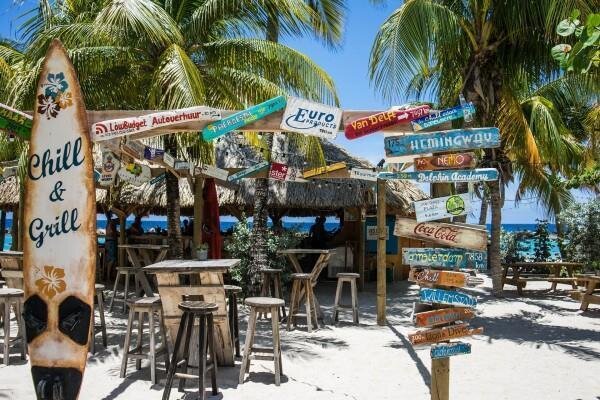 Strandbar mit Holzstühlen und Tischen unter Palmen, Wegweiser mit verschiedenen Ortsnamen und ein Surfbrett mit der Aufschrift Chill & Grill
