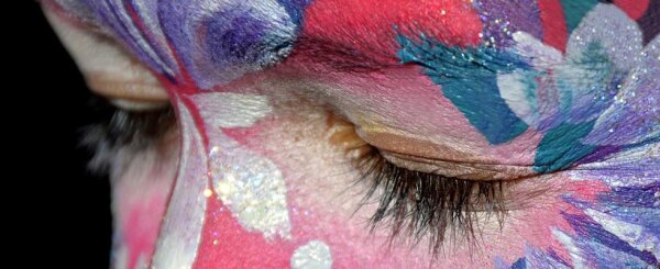 Nahaufnahme eines geschlossenen Auges mit langen Wimpern und buntem Gesichtsbemalung in Rosa, Weiß, Blau und Lila